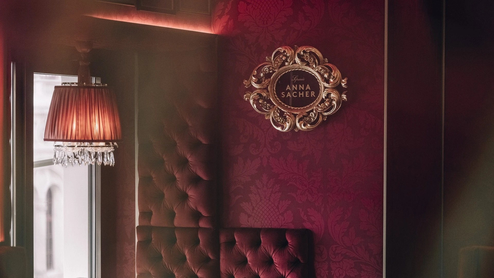 The interior of Café Anna Sacher in Vienna, with deep red tufted walls, an ornate gold-framed sign, and a crystal chandelier.