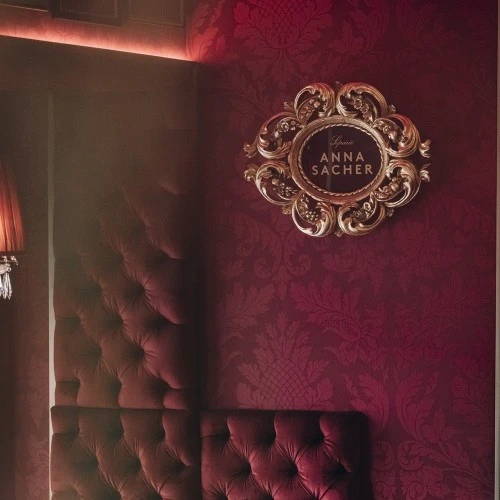 The interior of Café Anna Sacher in Vienna, with deep red tufted walls, an ornate gold-framed sign, and a crystal chandelier.