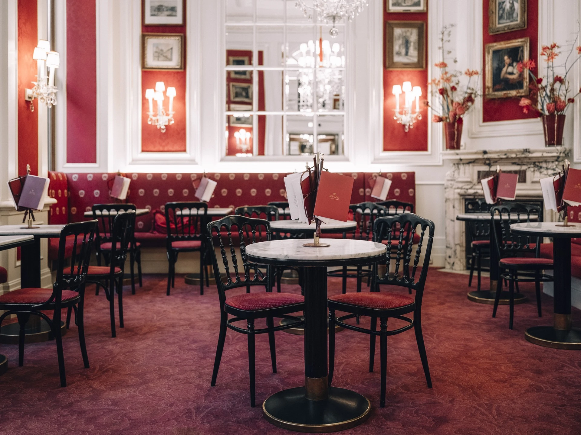 This image shows the grand interior of Café Sacher Wien, with its signature red and white palette, marble-topped tables, ornate chandeliers, and plush banquettes creating a timeless Viennese coffeehouse atmosphere full of elegance and history.