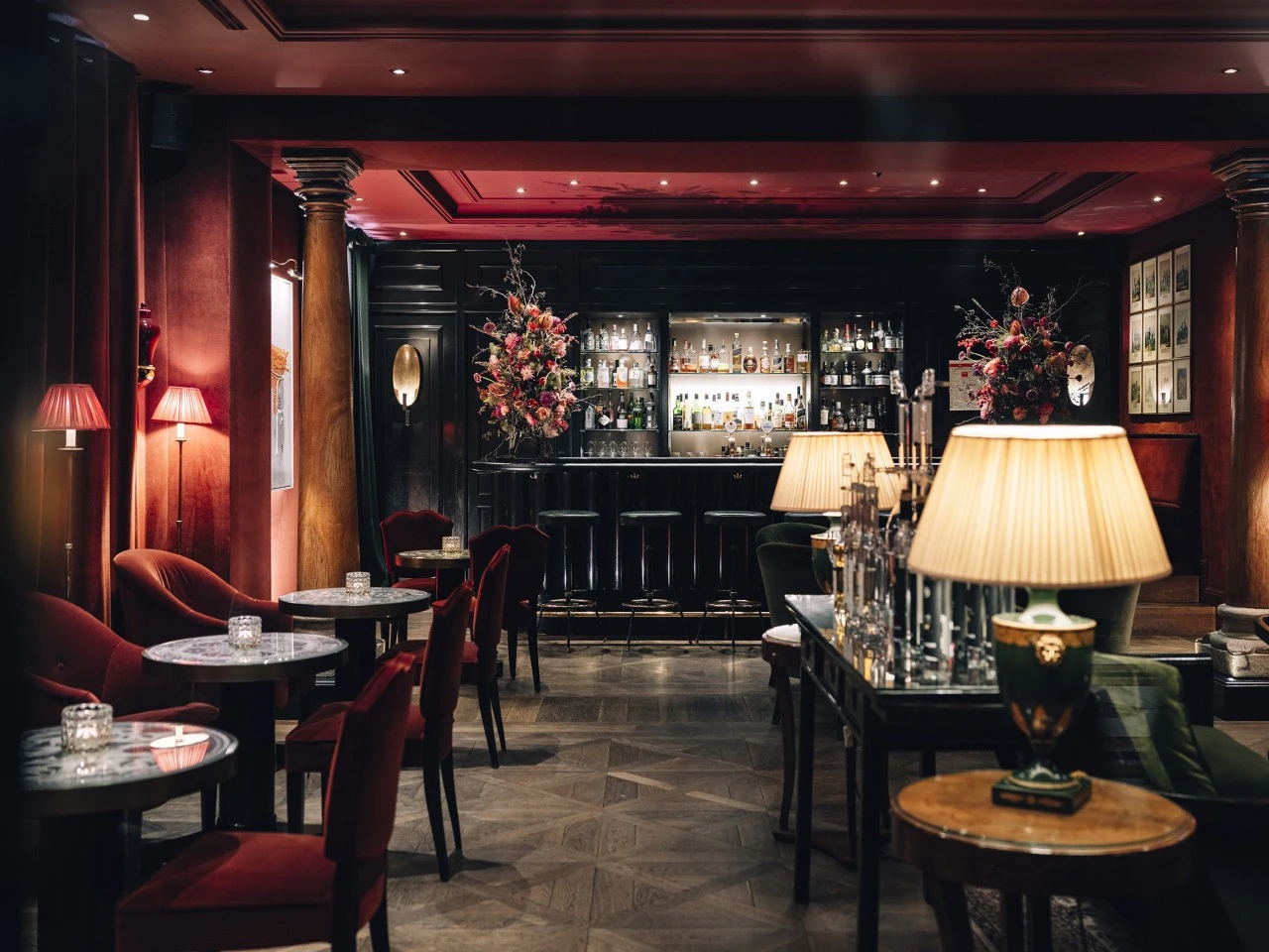 This image shows the lavish interior of the Sacher Bar at Hotel Sacher Salzburg, featuring deep red velvet furnishings, elegant marble-topped tables, dramatic floral arrangements, and a sophisticated bar illuminated by warm ambient lighting.