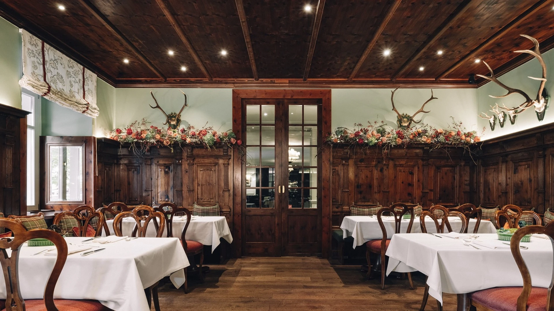 This wide-angle view of the Zirbelzimmer at Hotel Sacher Salzburg highlights its traditional alpine character, with intricately carved wooden paneling, antler displays, rich red seating, and floral garlands creating a warm and rustic yet refined dining ambiance.