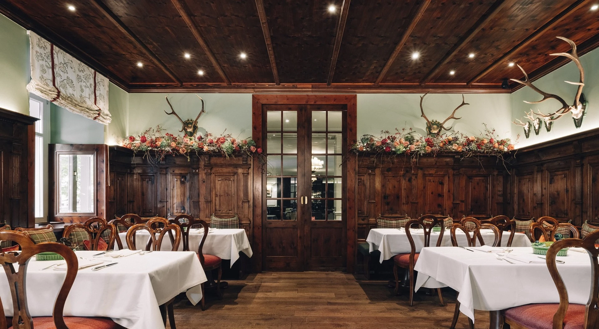 This wide-angle view of the Zirbelzimmer at Hotel Sacher Salzburg highlights its traditional alpine character, with intricately carved wooden paneling, antler displays, rich red seating, and floral garlands creating a warm and rustic yet refined dining ambiance.