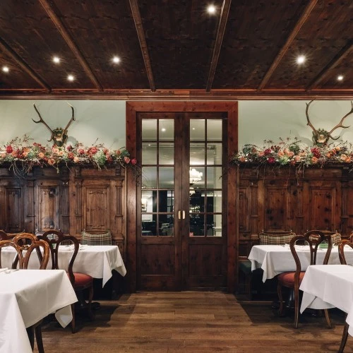 This wide-angle view of the Zirbelzimmer at Hotel Sacher Salzburg highlights its traditional alpine character, with intricately carved wooden paneling, antler displays, rich red seating, and floral garlands creating a warm and rustic yet refined dining ambiance.