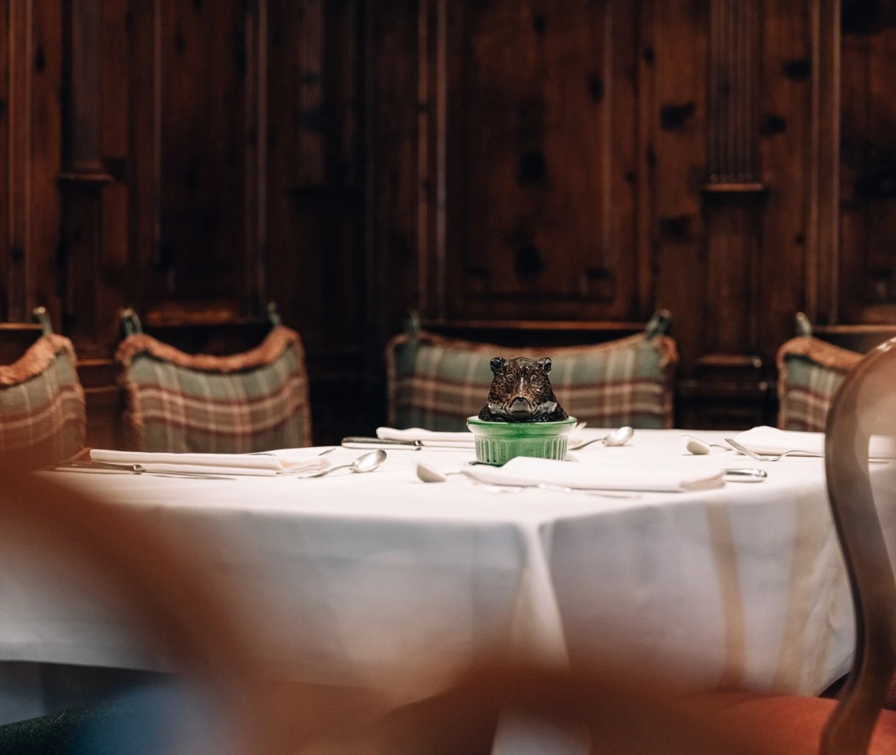 This close-up from the Zirbelzimmer at Hotel Sacher Salzburg highlights a table set with fine cutlery and napkins, centered around a charming green dish topped with a decorative ceramic boar’s head, all framed by rustic plaid cushions and dark wood paneling.
