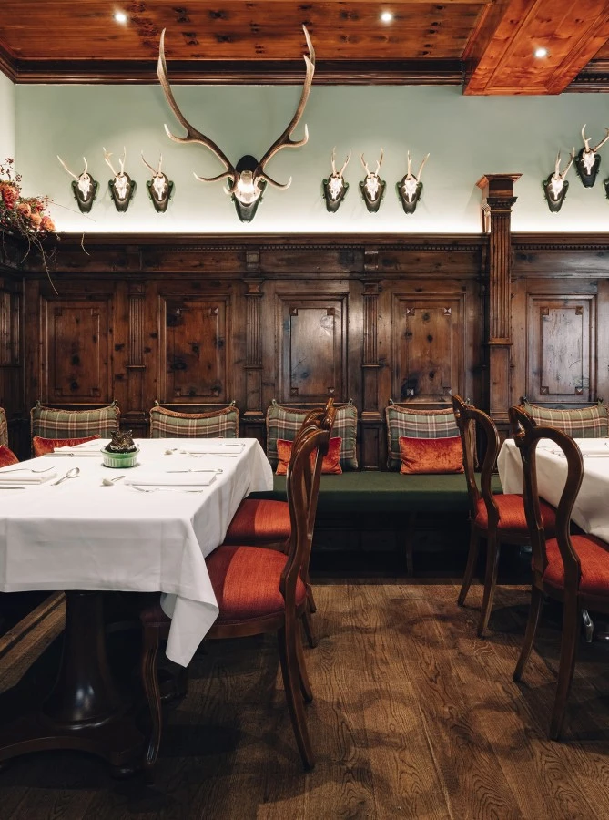 This image captures the cozy, alpine charm of the Zirbelzimmer at Hotel Sacher Salzburg, with its rich wood-paneled walls, traditional plaid upholstery, antler mounts, floral garlands, and elegant tables set with white linens for an authentic Austrian dining experience.