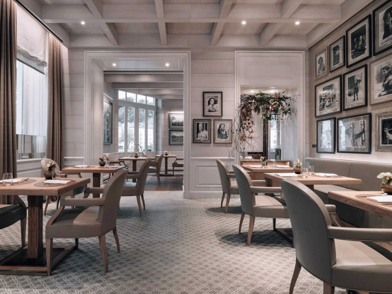 This image offers a bright and elegant view of the Sacher Grill at Hotel Sacher Salzburg, with natural light streaming through large windows, soft-toned furnishings, artistic floral decor, and a gallery wall of celebrity portraits creating a stylish and inviting dining ambiance.