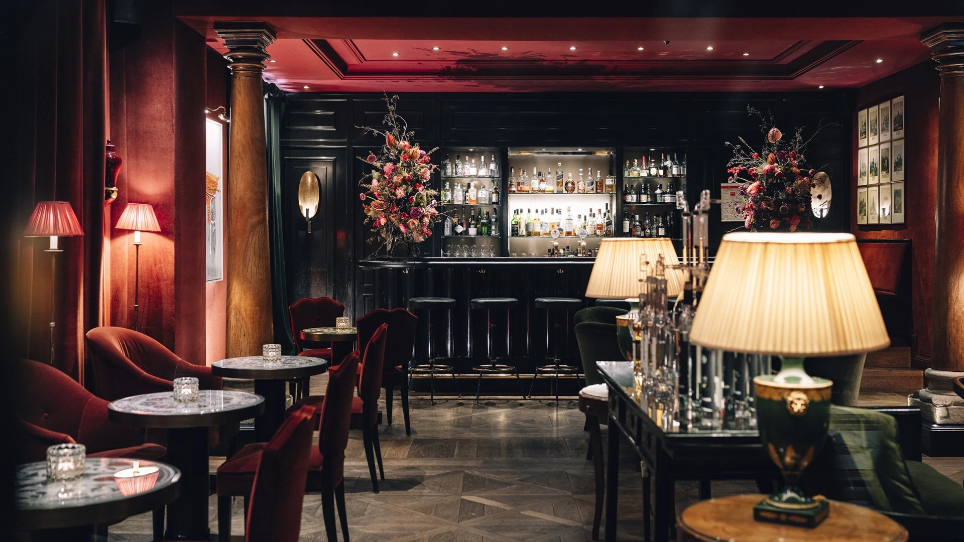 This wide-angle view of the Sacher Bar at Hotel Sacher Salzburg reveals its opulent and intimate setting, with deep red velvet furnishings, elegant marble-topped tables, golden lamps, and a dramatic black bar framed by lush floral arrangements and warm lighting.