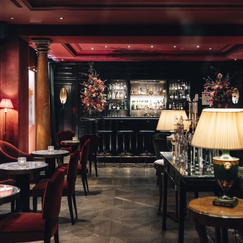 This wide-angle view of the Sacher Bar at Hotel Sacher Salzburg reveals its opulent and intimate setting, with deep red velvet furnishings, elegant marble-topped tables, golden lamps, and a dramatic black bar framed by lush floral arrangements and warm lighting.