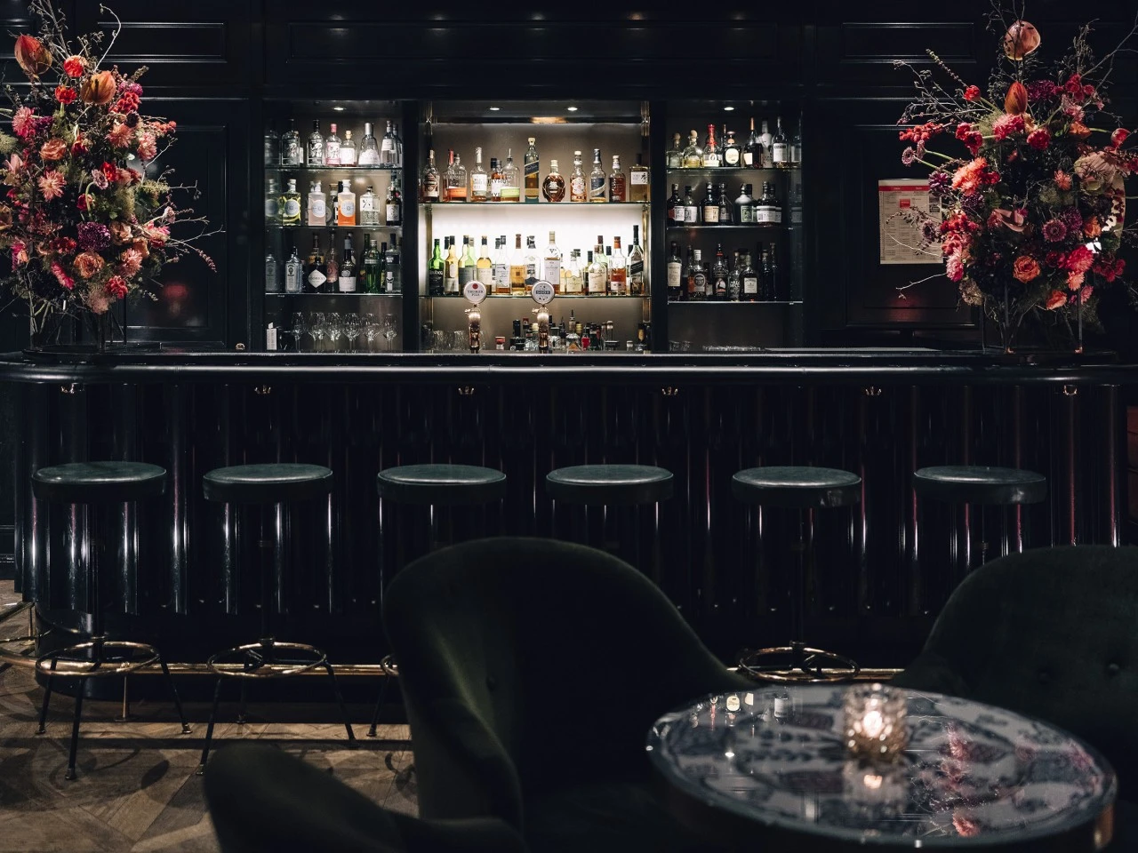 This image showcases the full view of the Sacher Bar at Hotel Sacher Salzburg, featuring a sleek black bar lined with velvet stools, an impressive selection of spirits, and dramatic floral arrangements that add elegance and vibrancy to the moody, luxurious ambiance.