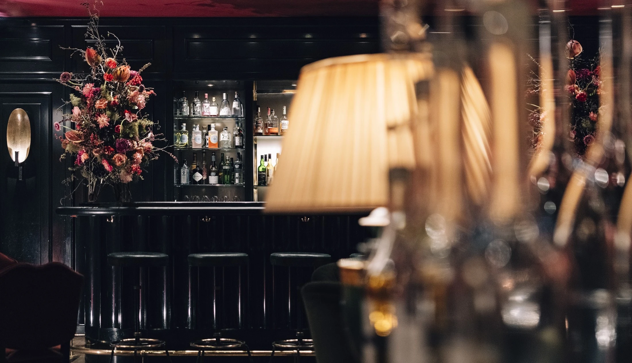 This image shows the elegant bar counter of the Sacher Bar at Hotel Sacher Salzburg, featuring a beautifully lit liquor display, dark lacquered wood, plush bar stools, and dramatic floral arrangements that add a luxurious, intimate touch.
