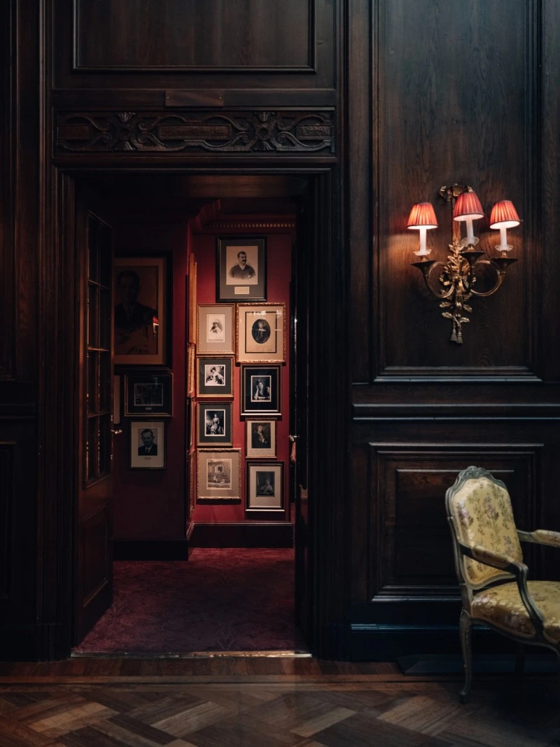 Dimly lit hallway at Hotel Sacher Vienna with vintage portraits on a red wall and an ornate chair beside wooden panelling.