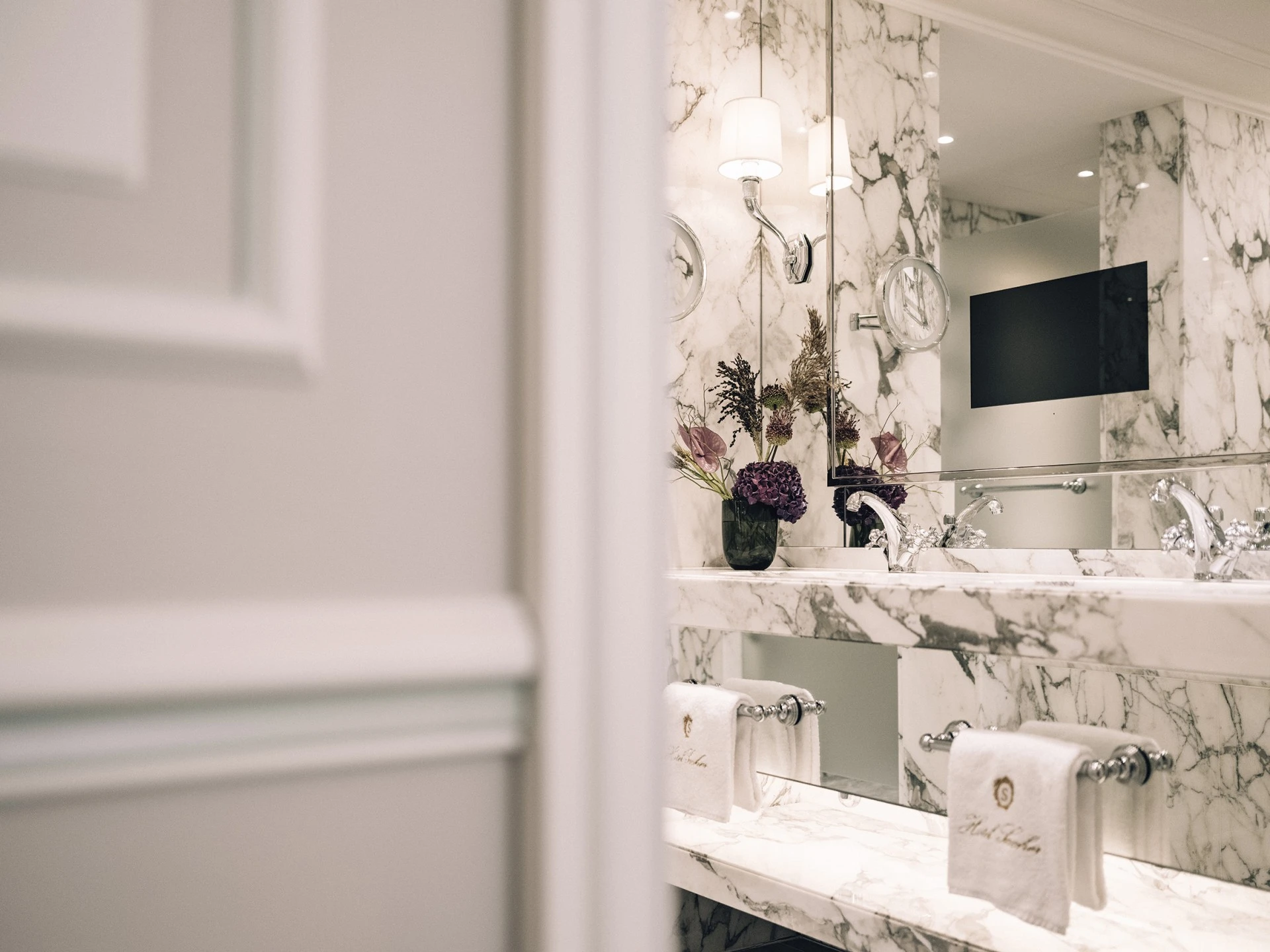 This image shows the luxurious marble bathroom of a deluxe room, featuring a double vanity with polished chrome fixtures, embroidered towels, and a striking floral arrangement reflected in a wide mirror beneath soft wall lighting.