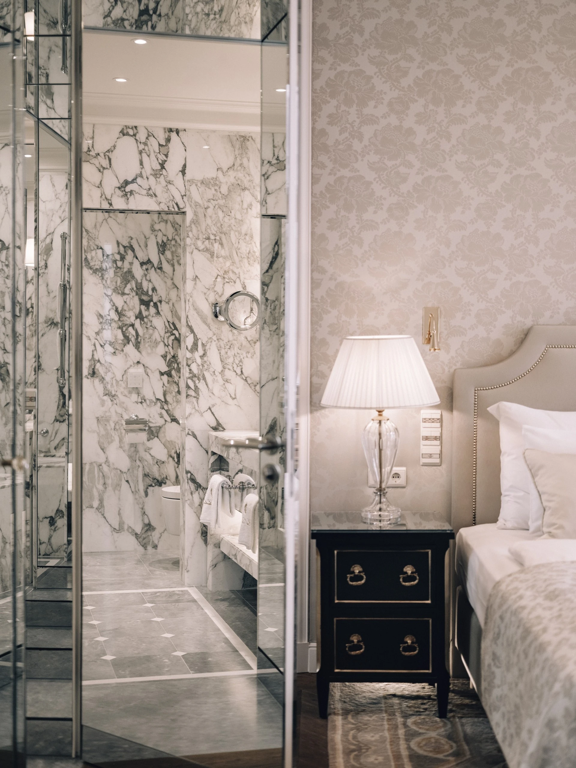 This image offers a seamless view from the junior suite’s elegant bedroom into the luxurious marble bathroom, blending classic charm with refined opulence through soft lighting, mirrored doors, and sophisticated detailing.