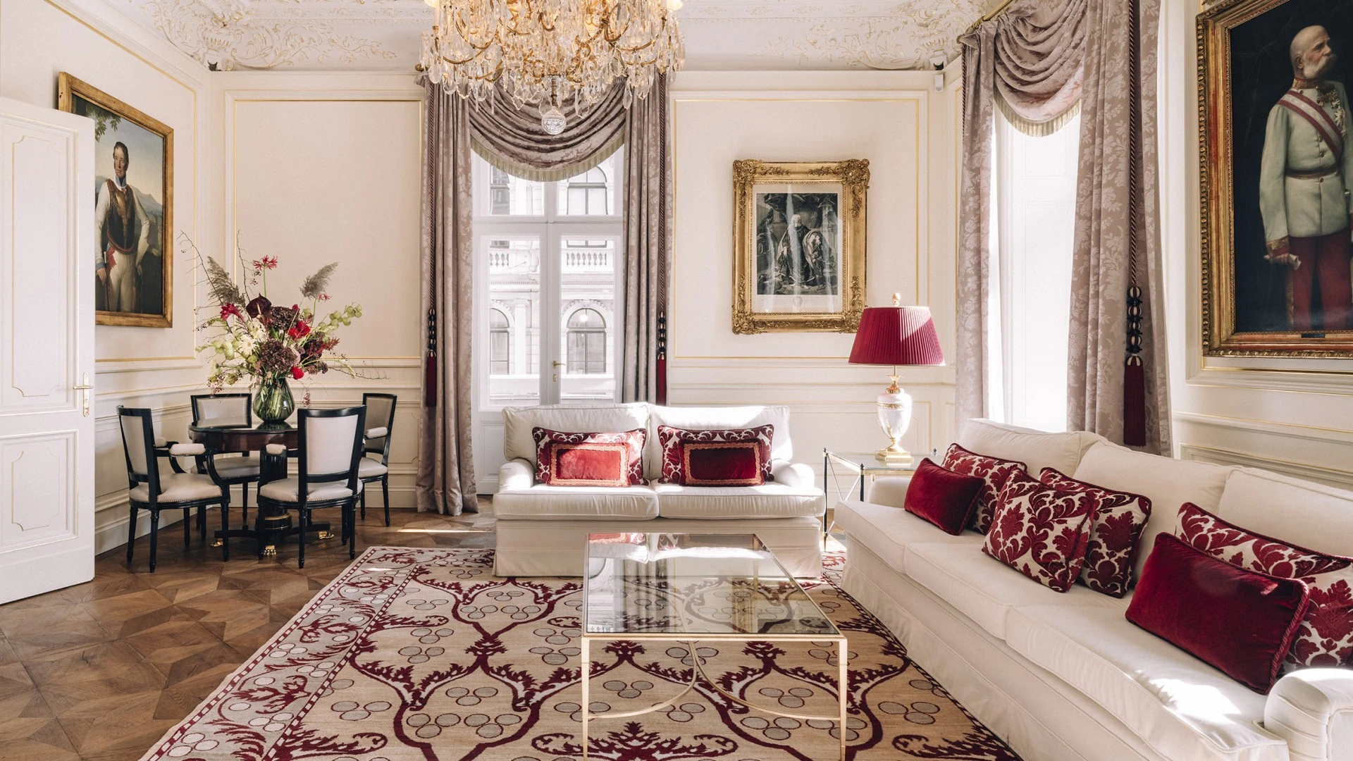 The image depicts the luxurious Philharmoniker Suite's living area, elegantly decorated with ornate ceilings, grand chandeliers, plush white sofas accented with red cushions, classical artwork, and refined floral arrangements, creating a sophisticated and opulent atmosphere.