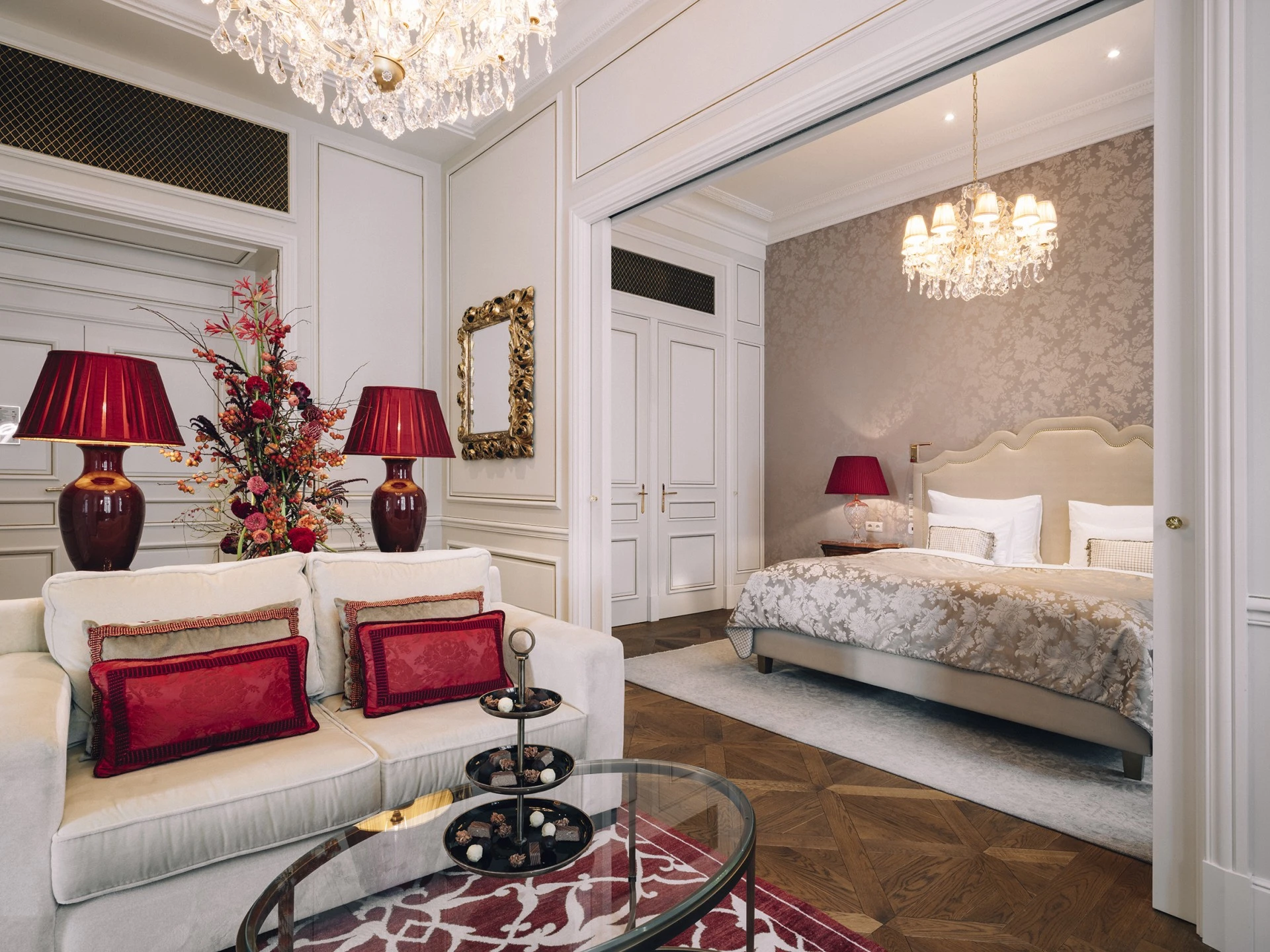 The luxurious Philharmoniker Suite, featuring an elegant bedroom and adjoining living area decorated with refined furnishings, red accents, floral arrangements, and crystal chandeliers, capturing a sophisticated Viennese charm.