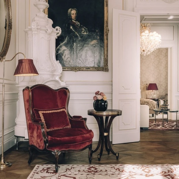 The image depicts an elegantly decorated room with a luxurious red velvet armchair, ornate white fireplace, classical artwork, and refined interior accents.