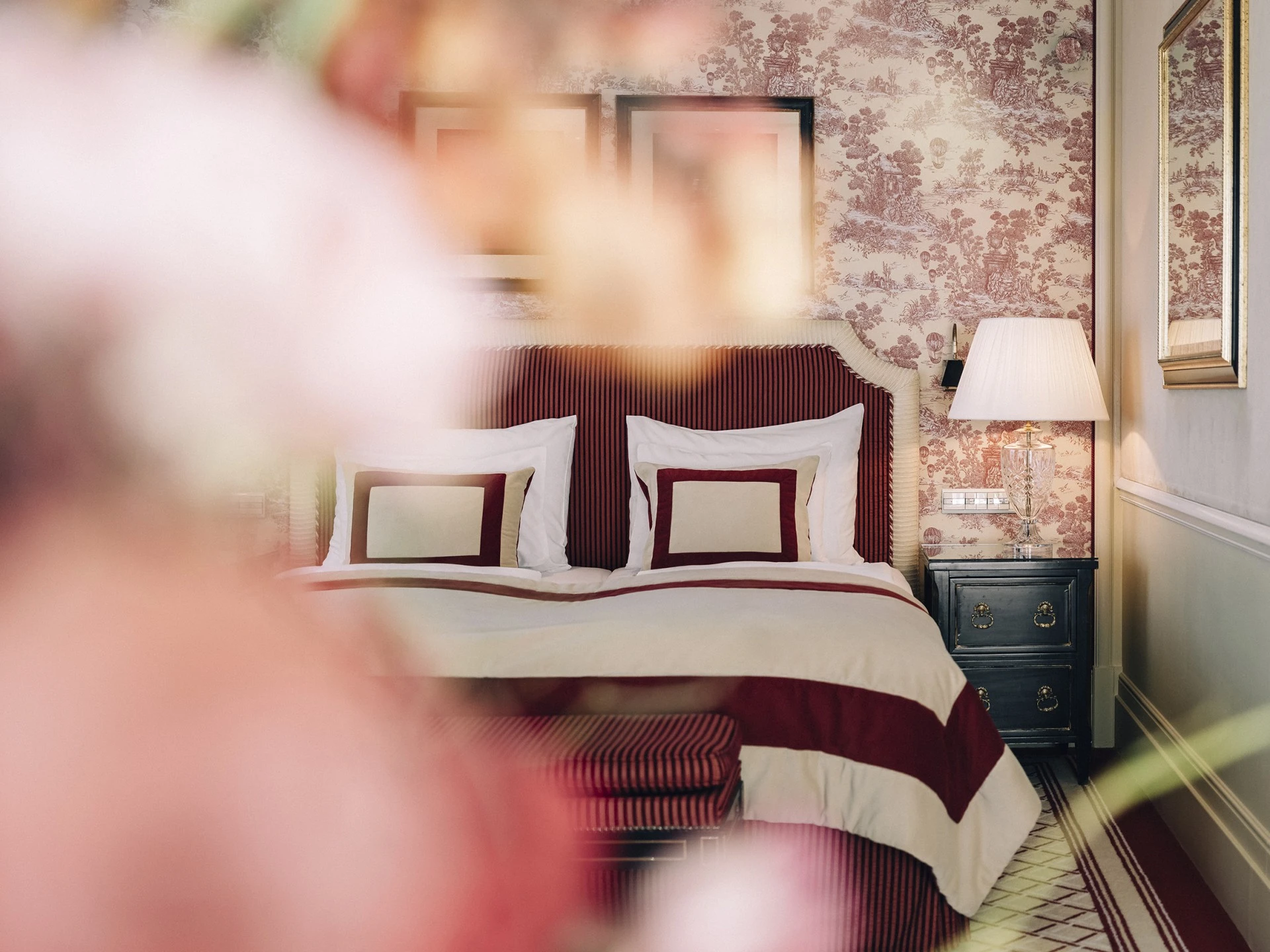 Bed with red and cream striped sheets, a nightstand with a lamp, and vintage floral wallpaper on the walls.