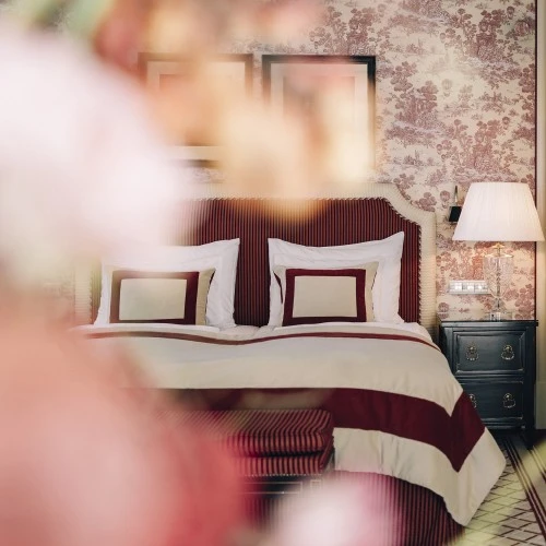Bed with red and cream striped sheets, a nightstand with a lamp, and vintage floral wallpaper on the walls.