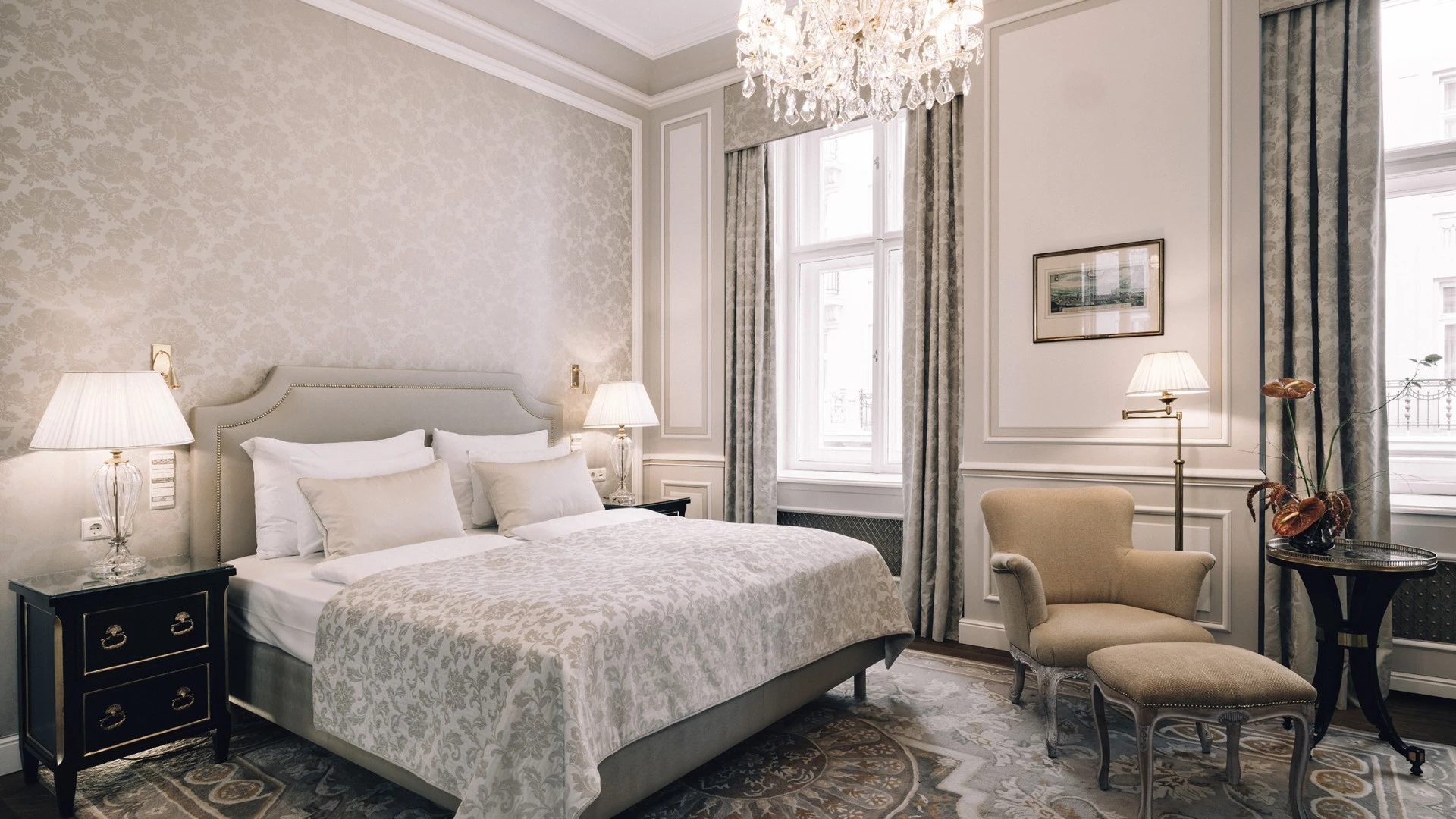 Junior Suite with an elegant and calm design. The room features a bed with soft lighting, a beige armchair with an ottoman, a floral rug, and large windows with light curtains. The space is decorated with a chandelier and a small side table with a plant, all giving the room a luxurious atmosphere.