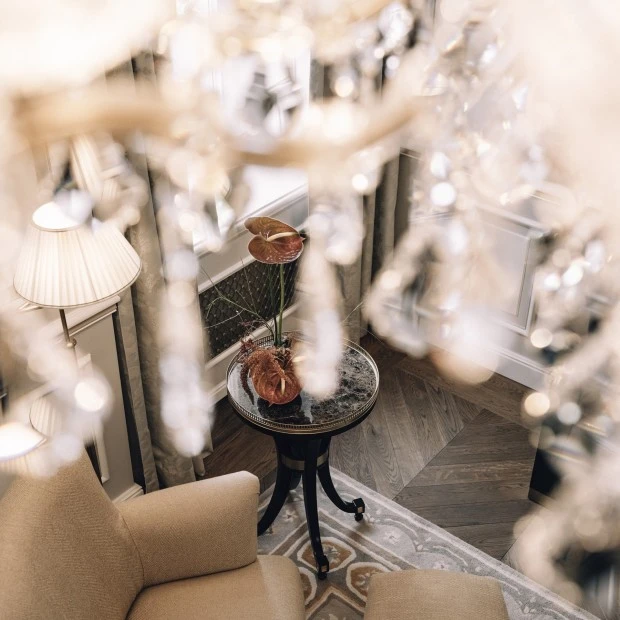 view of a luxurious sitting area within a suite. Two soft armchairs, a round table adorned with a plant, and a beautiful chandelier hanging above, partially blurred to create a soft, elegant effect.