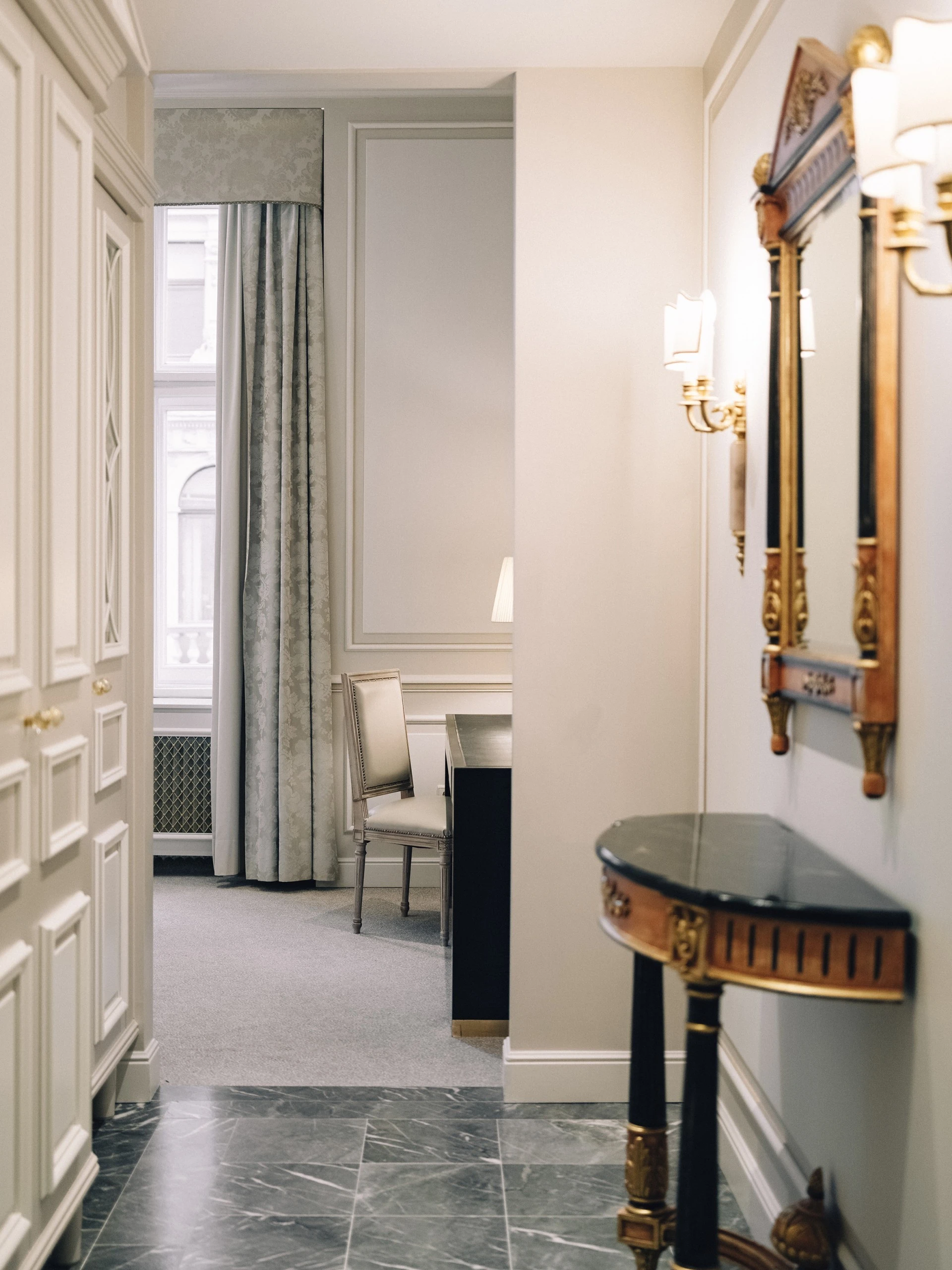 A hallway in a luxurious room with a marble floor, a small console table with a mirror above it, and a chair placed near a window with elegant curtains.