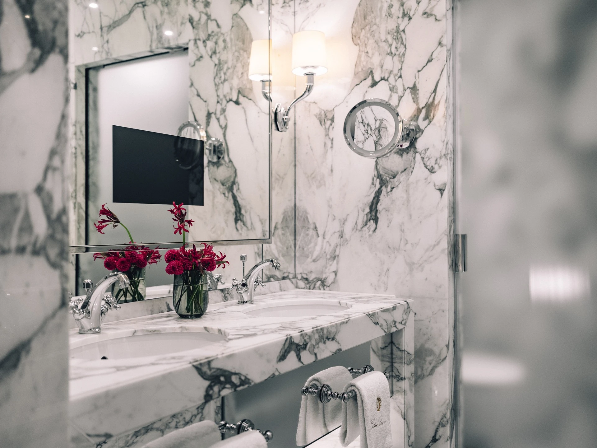 This is a luxurious bathroom at Hotel Sacher Wien, featuring marble walls, a pristine white sink, elegant faucets, and a decorative bouquet of red flowers on the countertop. There is also a modern mirror and a vanity light, adding to the sophisticated ambiance.