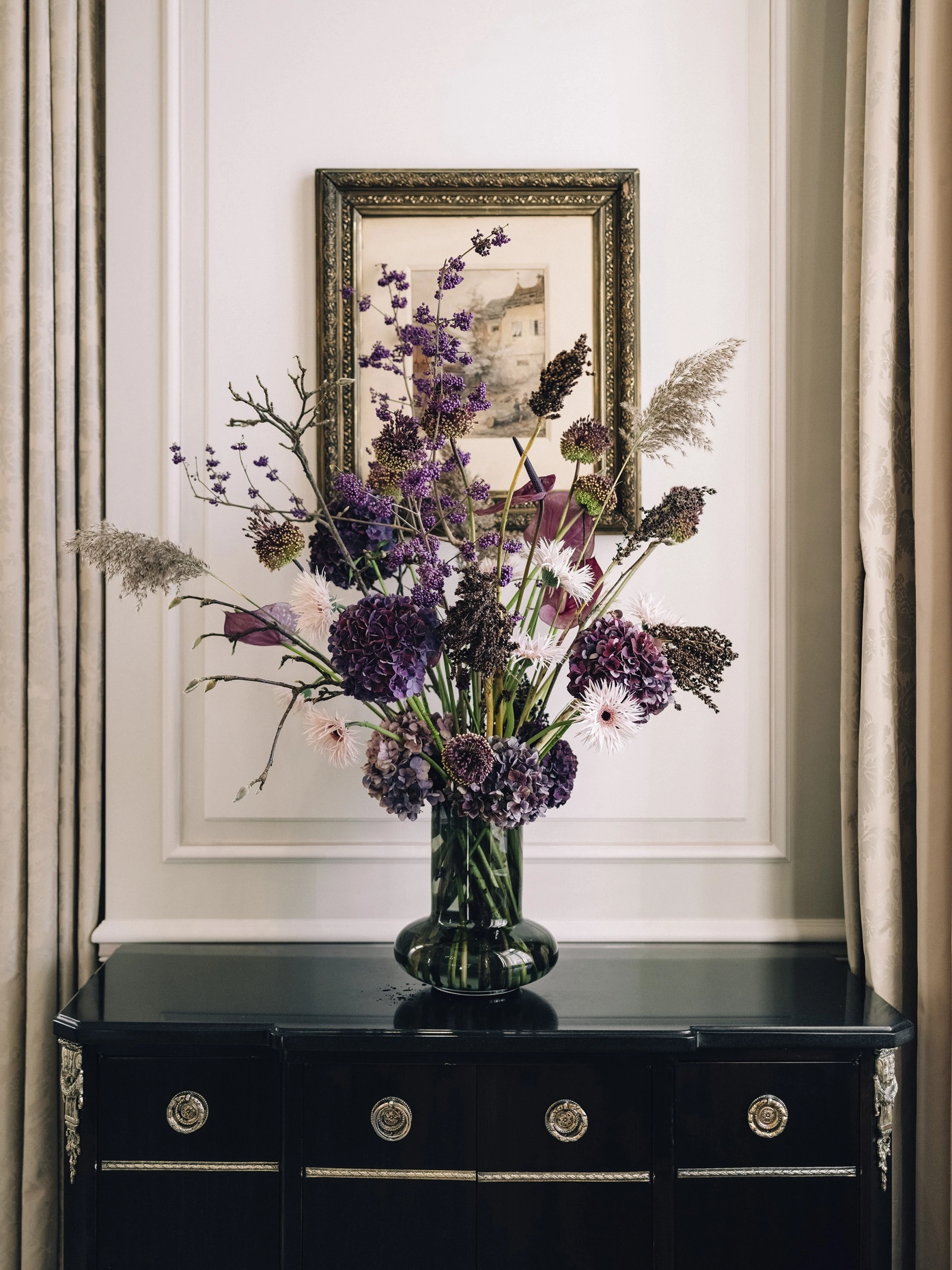 A large, elegant bouquet of purple, pink, and cream flowers arranged in a glass vase sits atop a glossy black cabinet with silver detailing, framed by beige curtains and a classic painting on the wall behind.