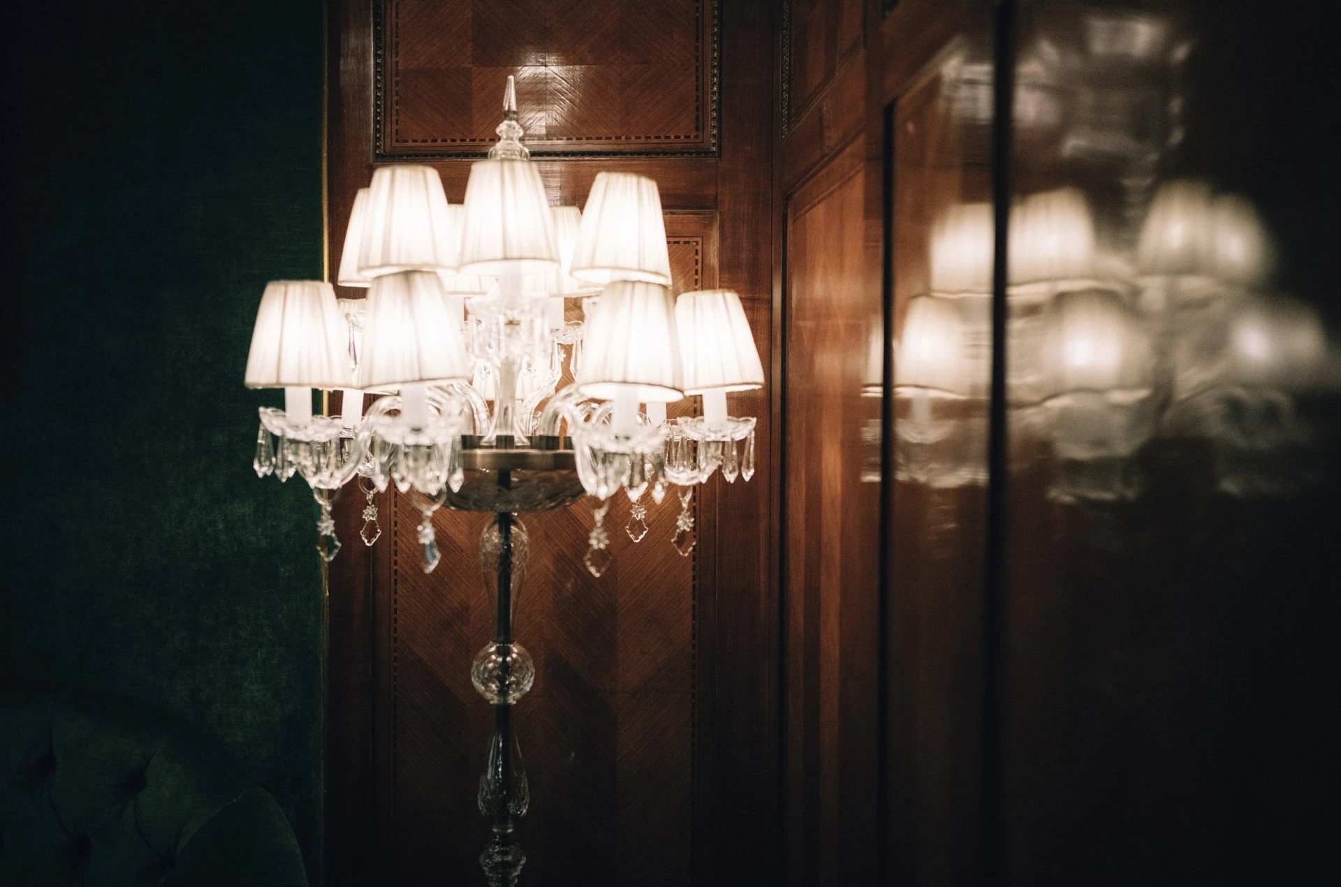 A crystal floor lamp with delicate white lampshades softly illuminates the richly paneled wood and deep green velvet walls, casting a warm, elegant glow in the timeless ambiance of Hotel Sacher Wien.