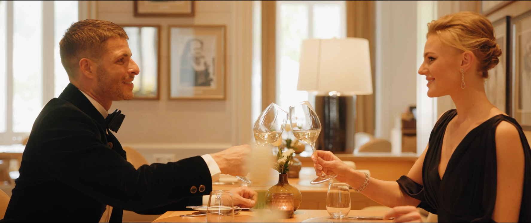A couple dressed in elegant evening wear clink glasses of white wine across a candlelit table, sharing a joyful toast in the refined ambiance of Hotel Sacher Salzburg.