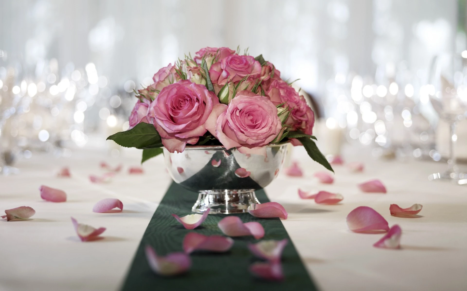 A silver bowl brimming with fresh pink roses sits at the center of a beautifully set table, scattered with delicate rose petals and glowing softly in elegant ambient light.