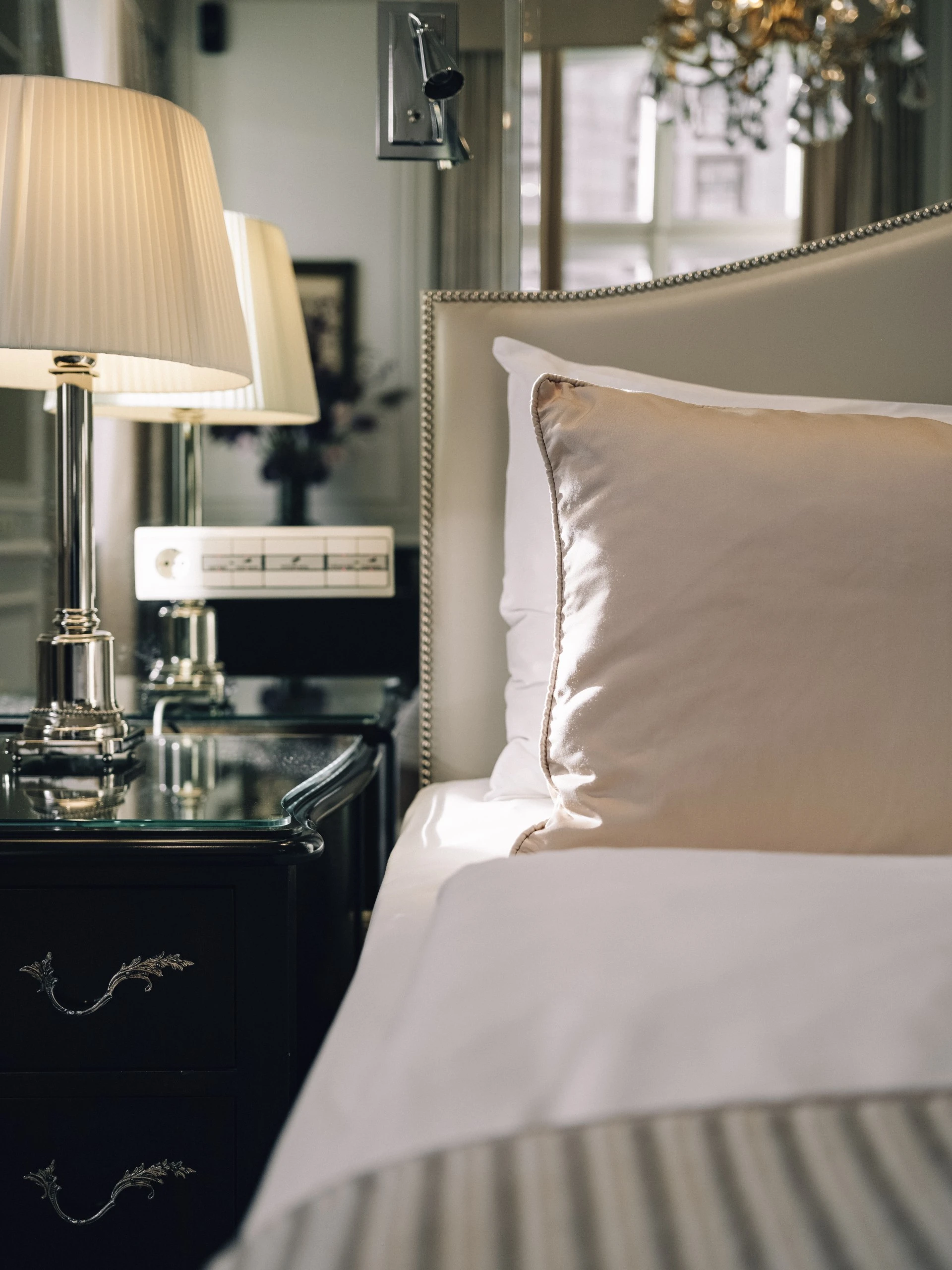 Soft sunlight bathes the edge of a perfectly made bed at Hotel Sacher Salzburg, highlighting a satin pillow, elegant nightstand, and classic lamp in a moment of quiet luxury.