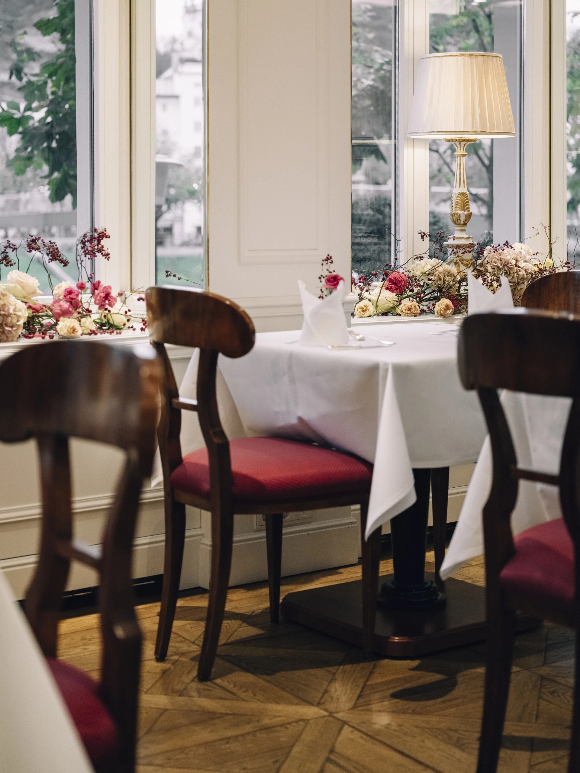 A beautifully set table with red-upholstered chairs and elegant floral arrangements sits by the window in the Salzachzimmer at Hotel Sacher Salzburg, offering a refined and intimate dining ambiance with views of the river.