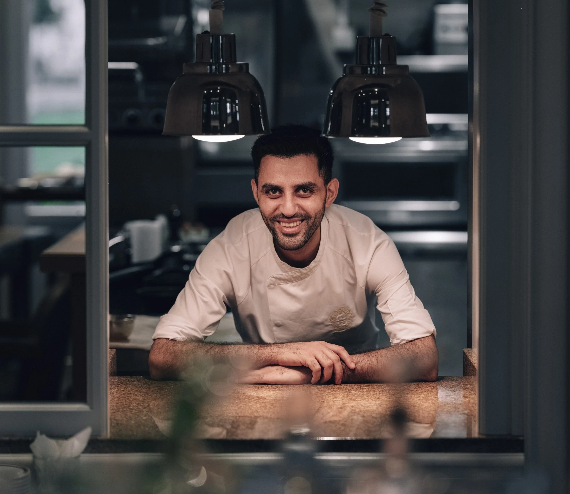 A smiling chef in a crisp white uniform leans proudly on the kitchen pass at Hotel Sacher Salzburg, warmly lit beneath hanging lamps, ready to share his culinary craft.