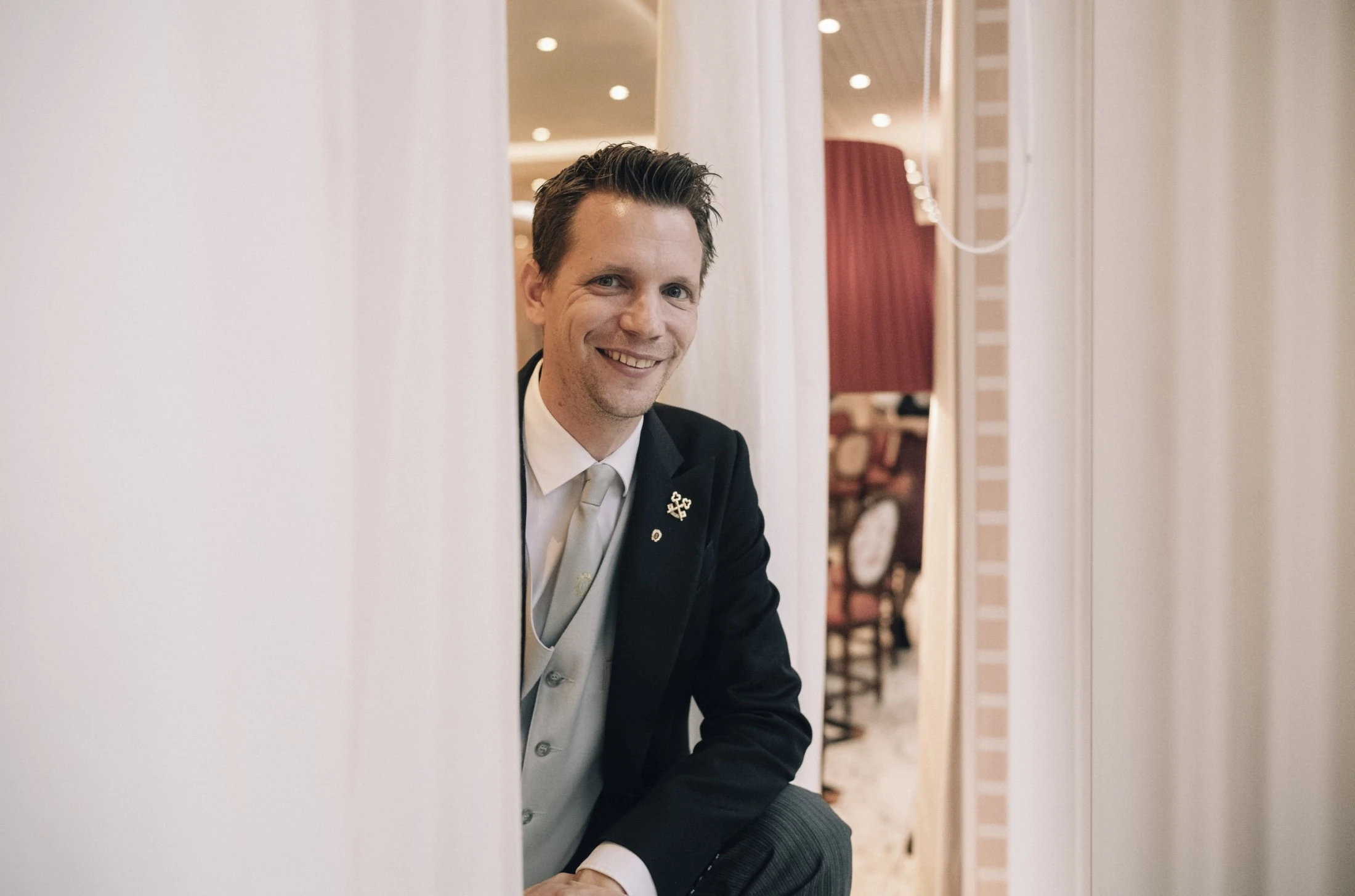 A smiling concierge at Hotel Sacher Salzburg, proudly wearing the golden keys of Les Clefs d'Or, peeks through elegant white curtains, ready to assist with impeccable service.