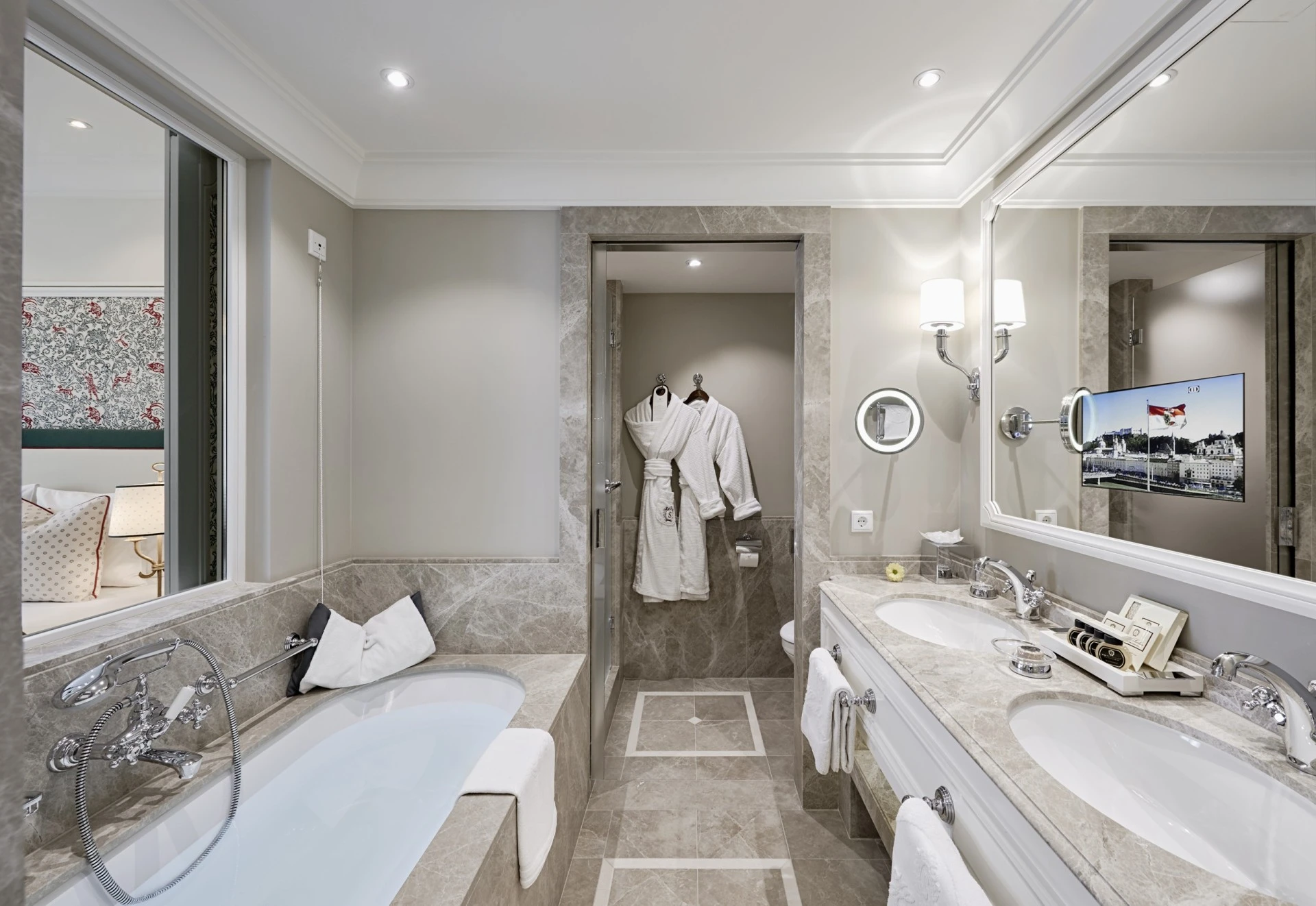 A luxurious marble bathroom at Hotel Sacher Salzburg features a deep soaking tub, plush robes, double sinks, and elegant amenities, offering a serene retreat with a glimpse into a cozy bedroom beyond.