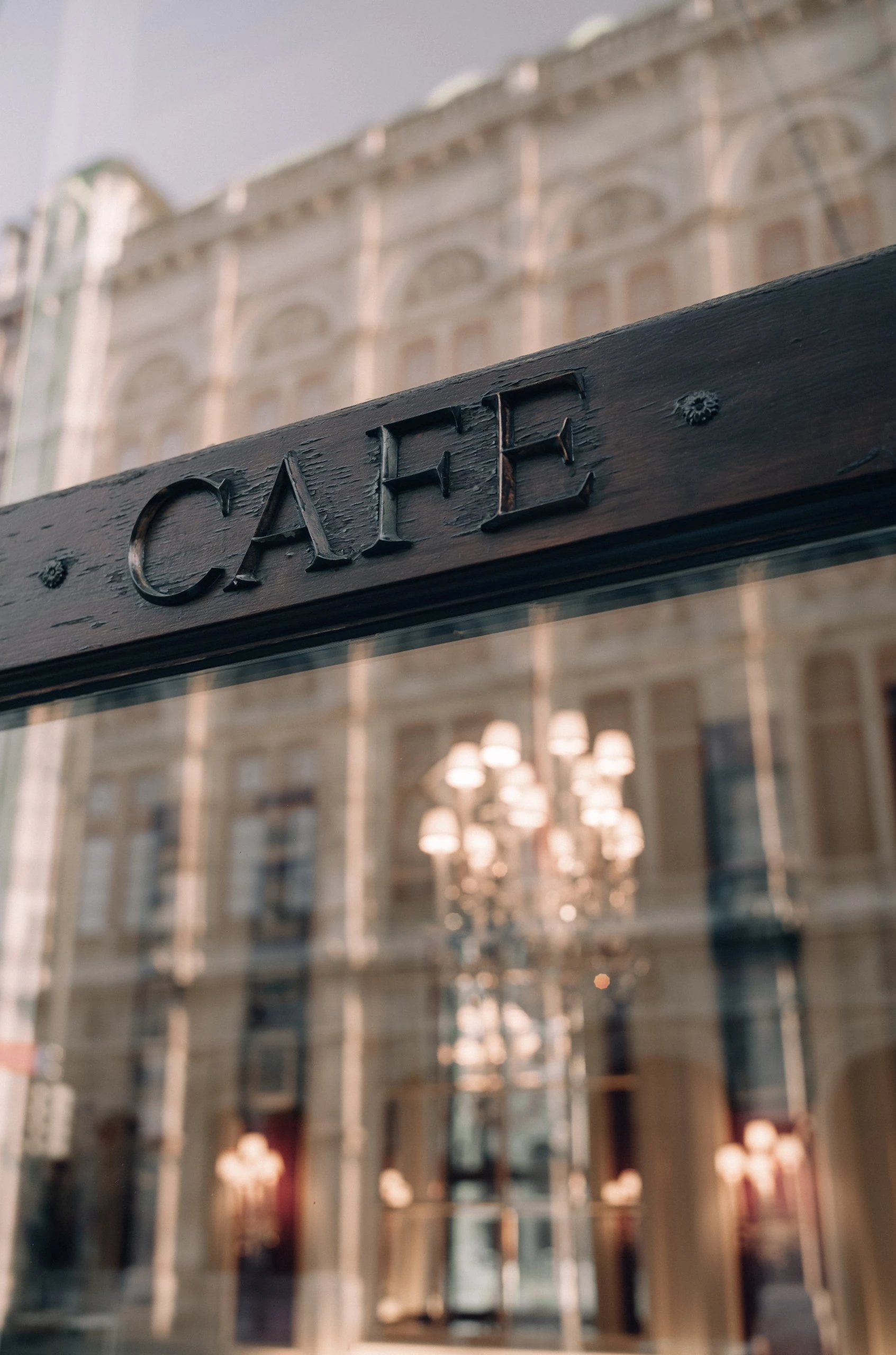 A close-up of a wooden sign reading “CAFE” reflects the elegant facade and chandeliers of the historic Café Sacher Vienna through a large window.