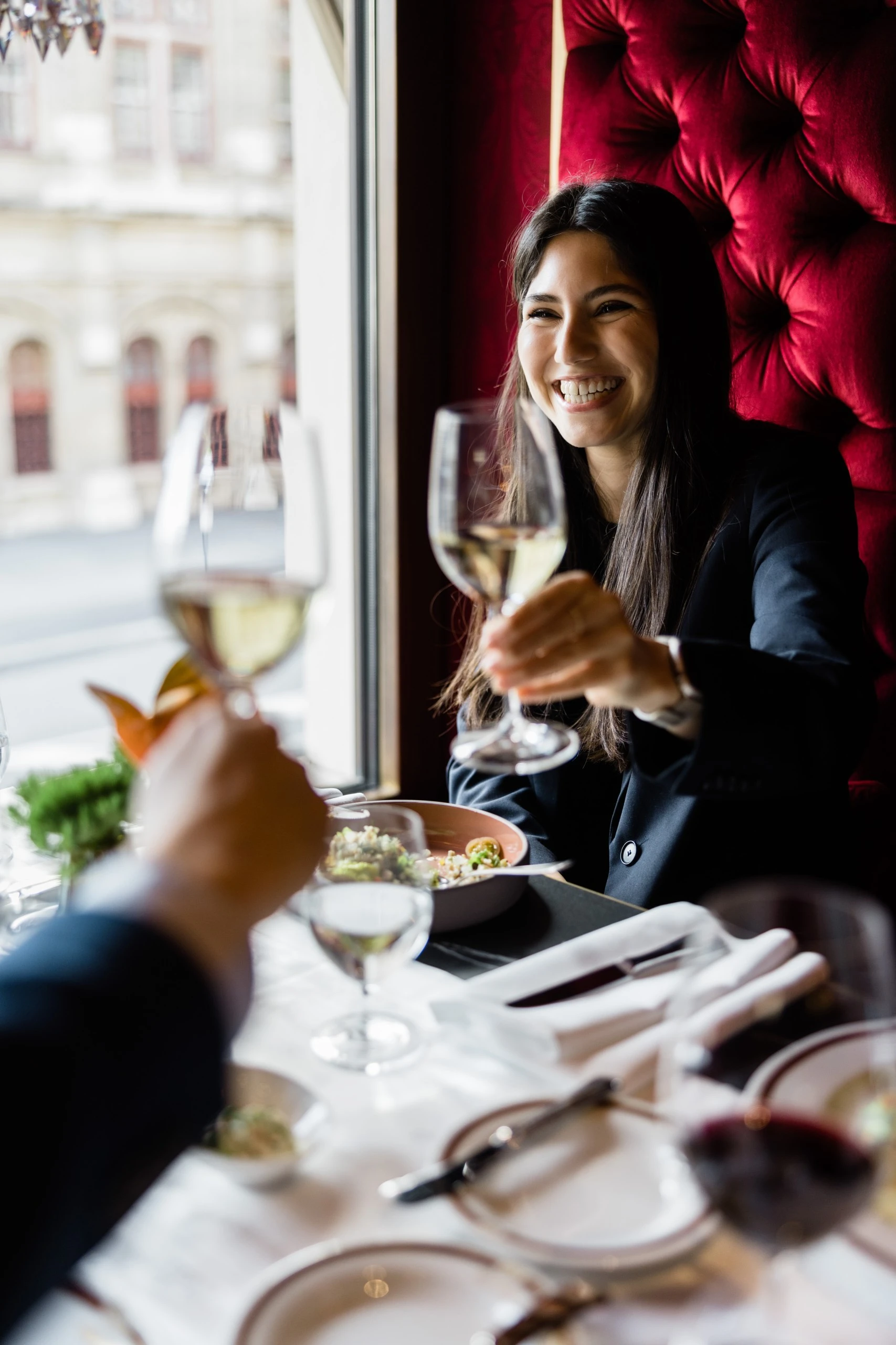 A smiling woman raises a glass of white wine in a toast across a beautifully set restaurant table by a window, exuding warmth and joy.