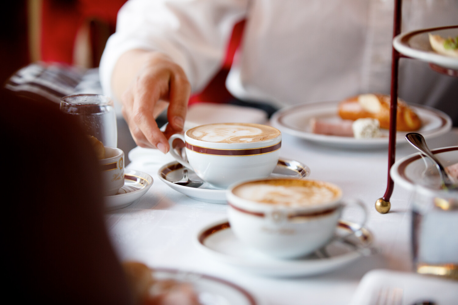 A hand reaches for a cappuccino with delicate latte art, served in elegant Hotel Sacher porcelain, as part of a refined breakfast setting with pastries and cold cuts on a white tablecloth.