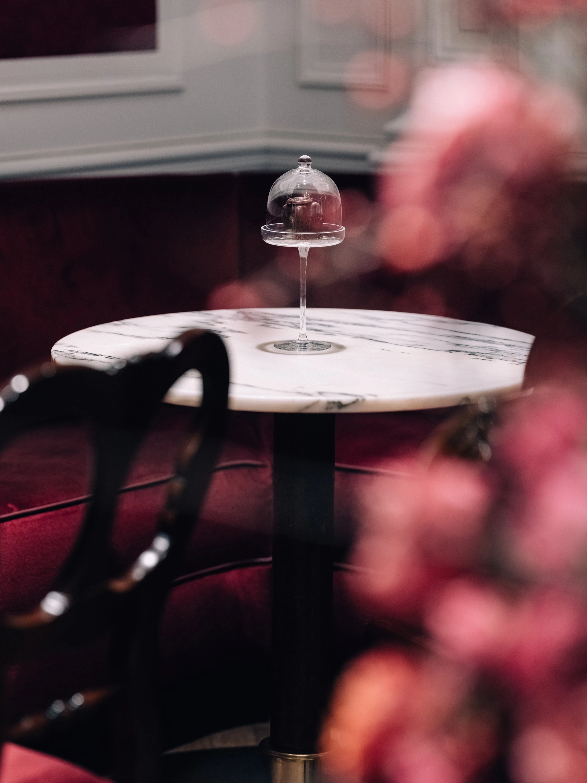 An Original Sacher Würfel sits elegantly under a glass dome on a tall stand, placed on a round marble table in a refined café setting with red velvet seating.