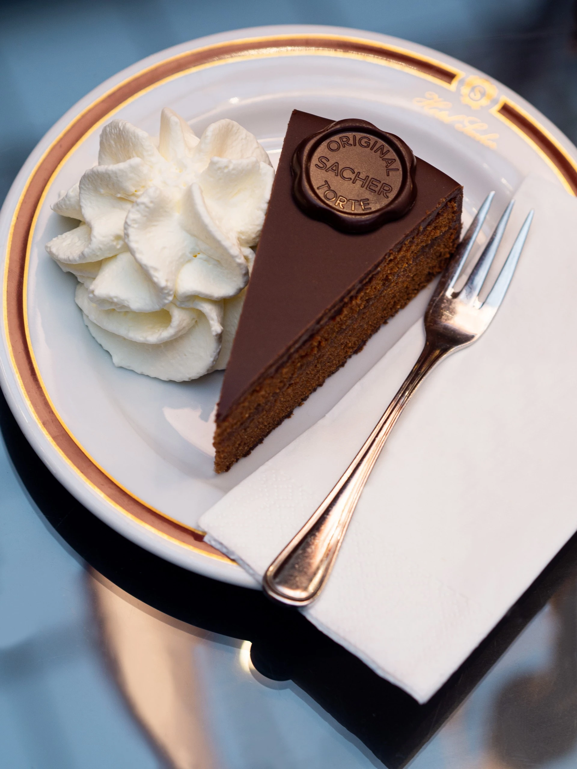 A slice of the Original Sacher-Torte with a glossy chocolate glaze and signature seal is served on elegant Hotel Sacher china alongside a swirl of whipped cream and a silver fork.