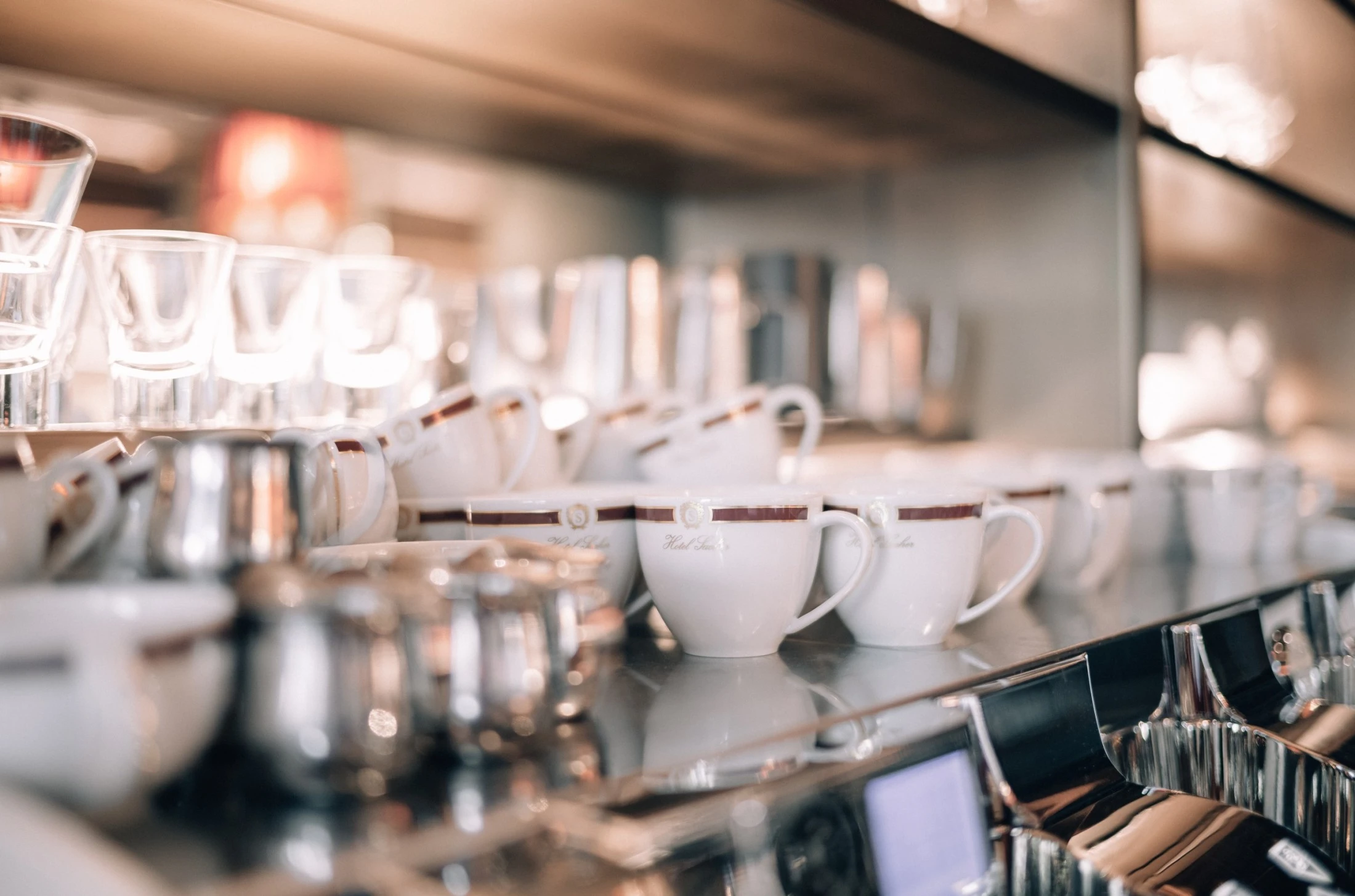 The image shows a close-up of an elegant coffee station with neatly arranged white porcelain cups bearing the “Hotel Sacher” logo, surrounded by gleaming metal milk jugs and glassware, all reflecting the soft ambient light of a refined café setting.