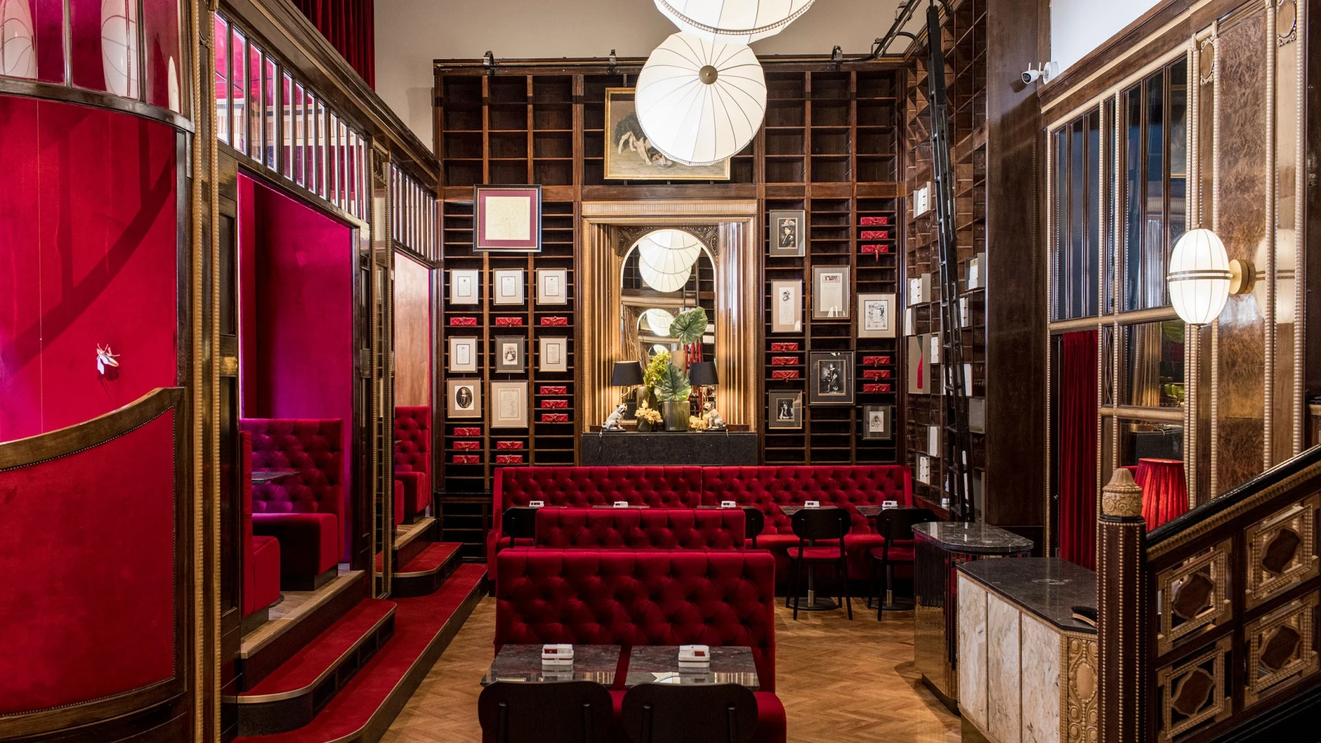 This image shows the richly decorated interior of Café Sacher in Trieste, featuring plush red velvet seating, gold and dark wood detailing, walls lined with portraits and framed documents—including tributes to Anna Sacher—and neatly stacked red boxes of the original Sachertorte, creating a refined and historic Viennese-style atmosphere.