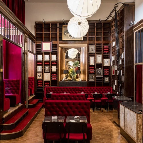 This image shows the richly decorated interior of Café Sacher in Trieste, featuring plush red velvet seating, gold and dark wood detailing, walls lined with portraits and framed documents—including tributes to Anna Sacher—and neatly stacked red boxes of the original Sachertorte, creating a refined and historic Viennese-style atmosphere.
