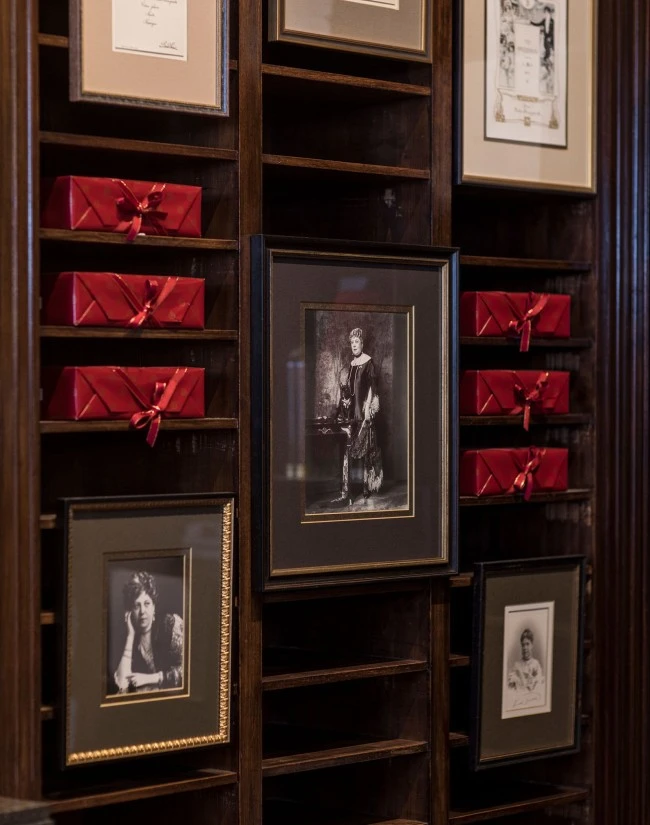 The image shows a dark wooden display wall featuring vintage black-and-white portraits of Anna Sacher, an elegantly dressed woman known for her role in Viennese hospitality, along with framed documents and several red ribboned boxes containing the original Sachertorte, the iconic Austrian chocolate cake.