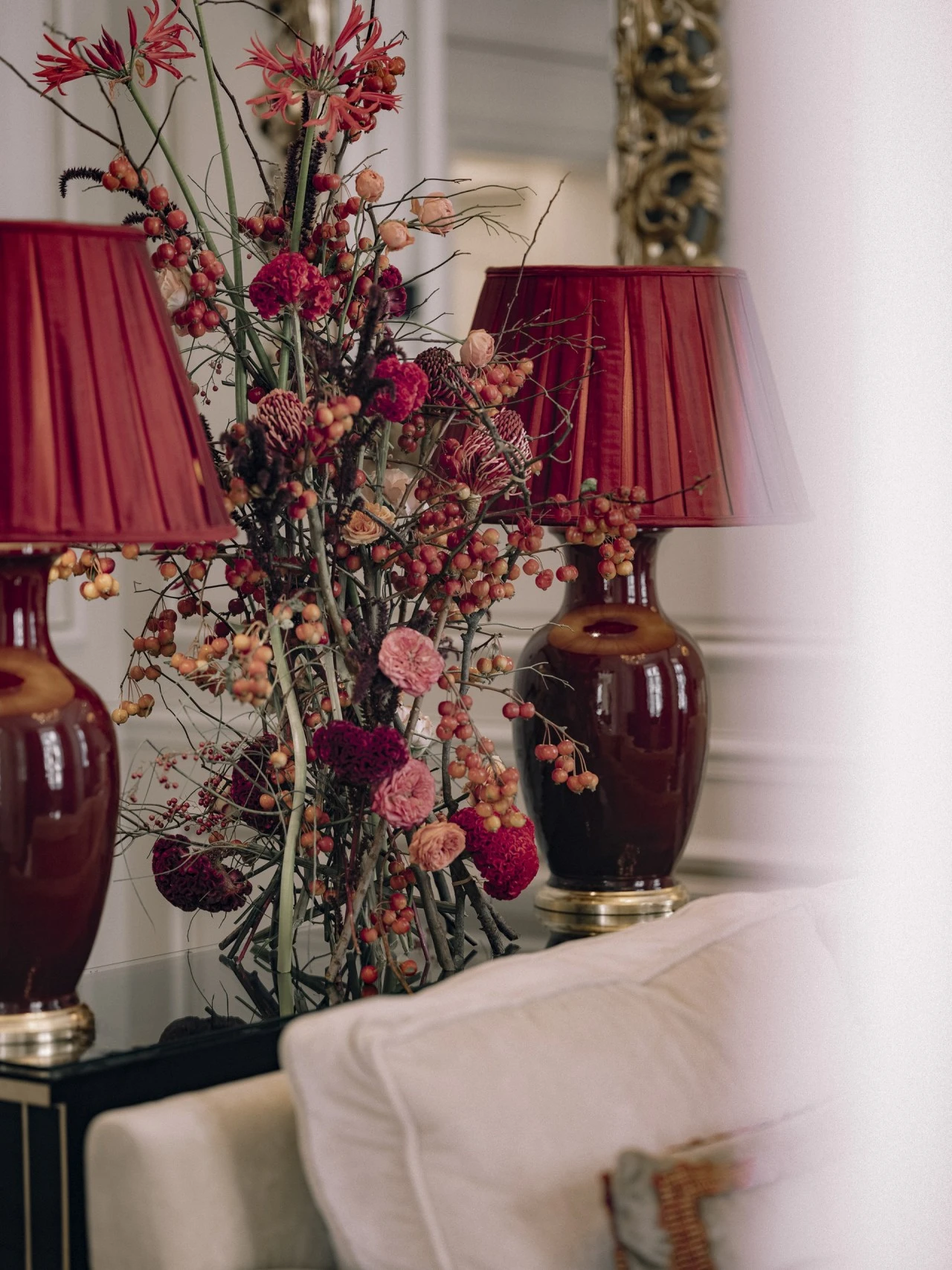 This image shows an elegant floral arrangement in rich shades of red and pink set between two deep red ceramic lamps with matching pleated shades, placed on a glossy black console behind a cream sofa in the refined setting of Hotel Sacher Vienna.