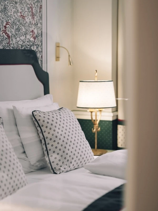 This image shows a stylish bed at Hotel Sacher Salzburg with crisp white linens and patterned pillows, set against an ornate wallpaper with red and black details, warmly lit by a classic lamp and brass reading light.
