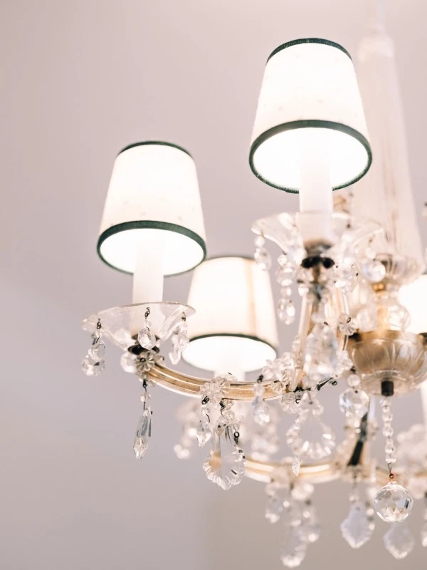 This image shows a close-up of an elegant crystal chandelier with delicate lampshades and sparkling hanging ornaments, glowing warmly against a soft, neutral ceiling background.