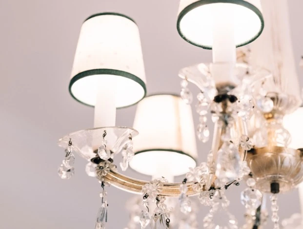 Zimmer-&-Suite-Salzburg_V1_Web_80 (c) Hotel Sacher This image shows a close-up of an elegant crystal chandelier with delicate lampshades and sparkling hanging ornaments, glowing warmly against a soft, neutral ceiling background.