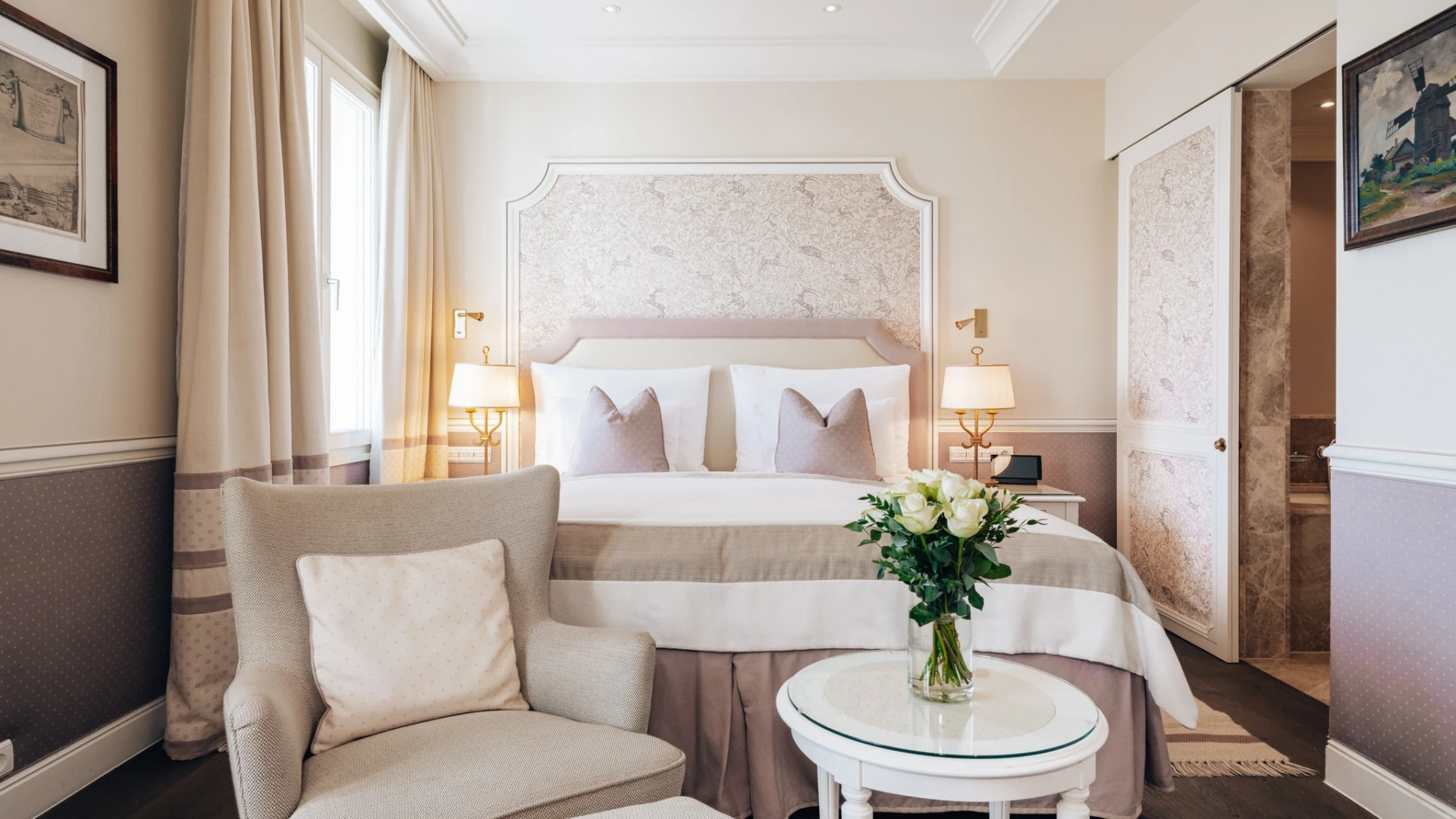 The image shows a cozy, elegant bedroom with soft beige and lavender tones, featuring a neatly made bed with decorative pillows, a plush armchair, a small round table with a vase of white roses, and warm lighting from bedside lamps.