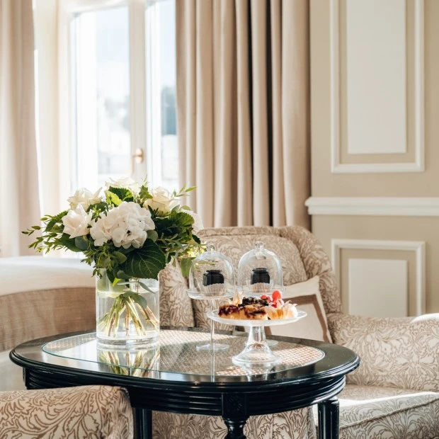 In a softly sunlit corner of a luxurious room, a round black table holds a vase of fresh white flowers and elegant pastries under glass domes, nestled between two patterned armchairs near tall curtained windows.